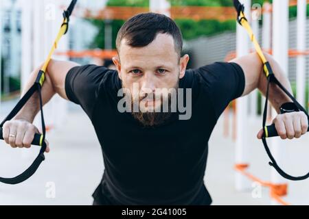 Konzentrierte Brünette mit einem Bart ist im Freien engagiert. Der Mann ist in Crossfit verwickelt, führt beharrlich Übungen durch, schaut auf die Kamera Stockfoto