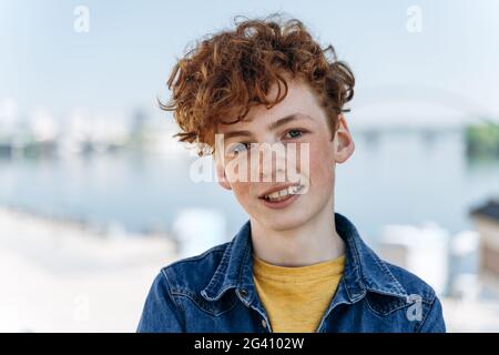 Hübscher, lächelnder Junge mit roten Haaren, der vor dem Hintergrund der Stadt posiert. Teenager, der vor dem Hintergrund einer geschäftigen Stadt posiert Stockfoto