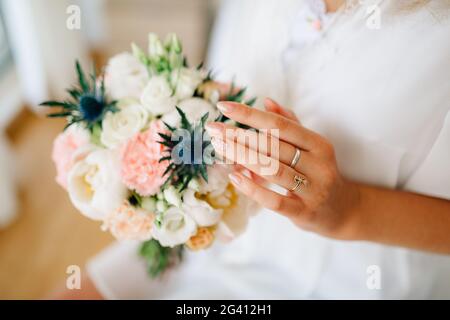 Die Braut hält sich in der Hand und berührt sanft ein Bouquet mit Pfingstrosen, Rosen, Lisianthus und Eryngien Stockfoto