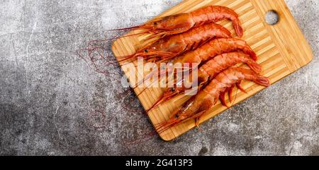 Argentinische rohe rote Garnelen (Garnelen) mit Köpfen auf einem rechteckigen Holzbrett auf dunkelgrauem Grund. Draufsicht, flach liegend Stockfoto