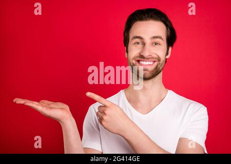 Foto von optimistischen Millennial braun Frisur Mann Haltepunkt leeren Raum tragen weißen T-Shirt isoliert auf rotem Hintergrund Stockfoto