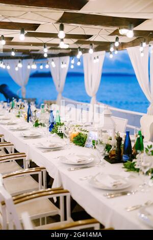 Hochzeitsessen Tischempfang. Ein langer Tisch, gegen das Meer, mit einer weißen Tischdecke, einer Girlande mit Glühbirnen Stockfoto