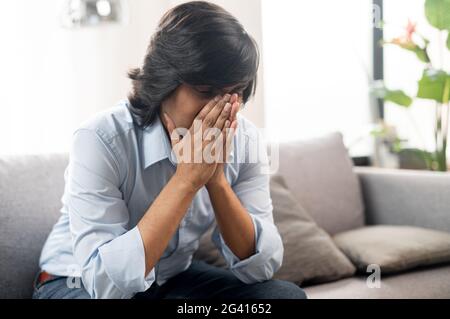 Unglücklicher junger inder, der auf dem Sofa sitzt, mit Händen bedeckt ins Gesicht sitzt und sich frustriert fühlt, müde und depressive Ostmann allein zu Hause, der an Migräne leidet Stockfoto