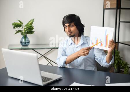 Selbstbewusster indischer Geschäftsmann, Lehrer oder Tutor, der ein Blatt des Dokuments hält und vorführt, mit virtuellen Meetings, Videokonferenzen, Online-Diskussionen über Verkaufsgraphen mit Kollegen Stockfoto