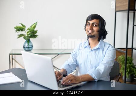 Porträt eines lächelnden indischen männlichen Mitarbeiters, der ein Headset verwendet und mit Kunden oder Kollegen spricht, eines hinduistischen Callcenter-Mitarbeiters mit einem Laptop, der am Schreibtisch im Büro sitzt Stockfoto