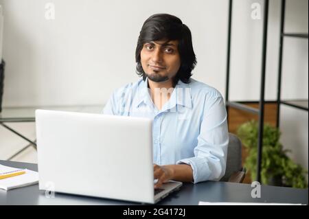 Ein gutaussehender inder in eleganter Freizeitkleidung, der im modernen Büro einen Laptop verwendet, ein Geschäftsmann aus dem Osten mit gemischtem Rennen blickt auf die Kamera. Technologie- und Lifestyle-Konzept Stockfoto