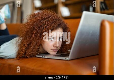 Traurig gelangweilte junge lockige rothaarige Frau, die sich vor dem geöffneten Laptop auf der Couch niederlegt und die Kamera anschaut, fühlt sich melanterisch und depressiv Stockfoto