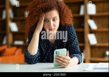 Besorgte rothaarige junge Freiberuflerin oder Studentin mit lockigen Haaren, die am Schreibtisch saß und das Smartphone mit rätselhaftem Gesichtsausdruck hielt, erhielt unerwartete, schockierte Nachrichten Stockfoto