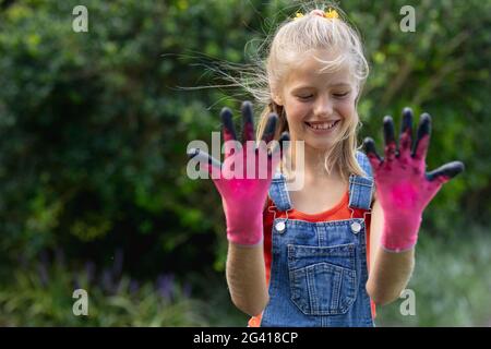 Lächelndes kaukasisches Mädchen im Garten, das die Hände in schmutzigen Händen hält Rosafarbene Gartenhandschuhe Stockfoto