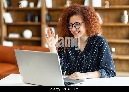 Optimistische Rothaarige lockige Geschäftsfrau, die Videoanruf auf dem Laptop macht, glückliche Freiberuflerin, die auf die Webcam schaut und winkt, begrüßt Online-Teilnehmer von Online-Meetings, virtuellen Konferenzen Stockfoto