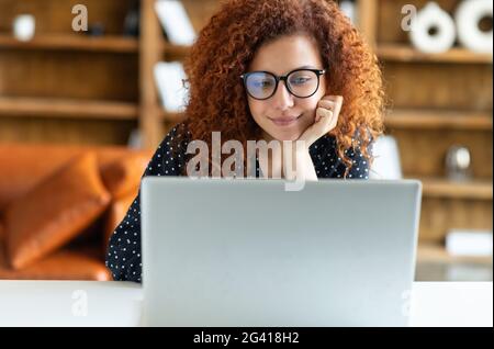 Porträt einer positiven rothaarigen lockigen, hübschen freiberuflichen Geschäftsfrau, die am Schreibtisch sitzt und auf einem Laptop tippt, mit einer Freundin chattet und an einem virtuellen Online-Projekt arbeitet Stockfoto