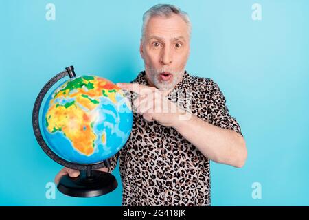 Foto von stauned schockiert alten Mann zeigen Finger Globus Touristenziel isoliert auf blauem Hintergrund Stockfoto