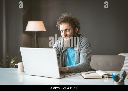 Der Texter arbeitet von zu Hause aus, um den Artikel zu schreiben. Ein junger Mann mit lockigen Haaren tippt auf einem Laptop. Arbeit zu Hause ist ein Koncep Stockfoto