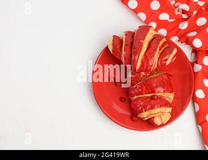 Zwei gebackene Croissants mit Kirschglasur auf einer roten Keramikplatte, weißer Tisch Stockfoto