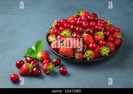Köstliche Kirschen und Erdbeeren auf einem schwarzen Teller auf blaugrauem Hintergrund. Seitenansicht. Sommerfrüchte und Beeren. Stockfoto