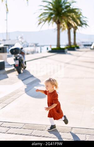 Nettes 18-Monate altes Mädchen in einem Terrakotta-Kleid und weiß Strumpfhosen laufen auf einem Bootssteg auf einem sonnigen Tag Stockfoto