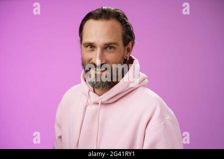 Charming verführerisch gut aussehend bärtig stilvolle Erwachsene männliche Modell Ohrring rosa Kapuzenpullover lächelnd erfreut Ausdruck Vertrauen Positivität Stockfoto