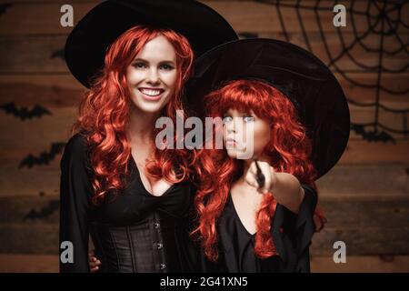 Schöne kaukasische Mutter und ihre Tochter mit langen roten Haaren in Hexenkostümen und Zauberstab feiern Halloween posiert Witz Stockfoto