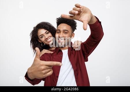 Glückliche junge afrikanische amerikanische Paar durch einen Finger Rahmen und lächelnd beim Stehen auf weißen isoliert. Stockfoto