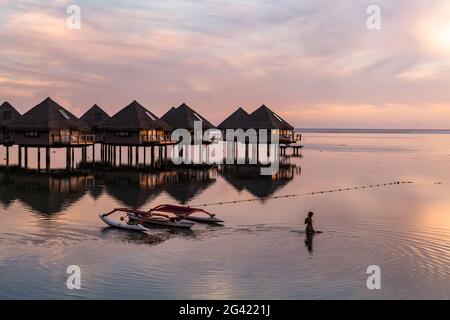 Silhouette einer Frau im Wasser neben dem Auslegerkanu und den Überwasser-Bungalows des Tahiti Ia Ora Beach Resort (verwaltet von Sofitel) bei Sonnenuntergang, Stockfoto