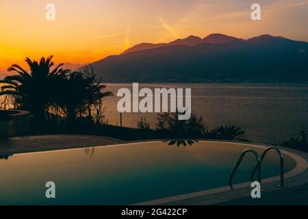 Der feurige Sonnenuntergang spiegelt sich im Swimmingpool wider, vor der Kulisse des Meeres, der Berge und der Palmen. Stockfoto