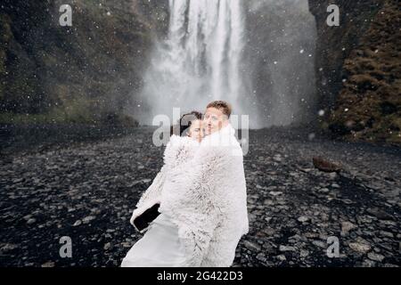 Hochzeitspaar in der Nähe des Skogafoss Wasserfalls. Die Braut und der Bräutigam waren mit einer Wolldecke bedeckt, wo sie umarmten. Schnee fällt, fallen Stockfoto