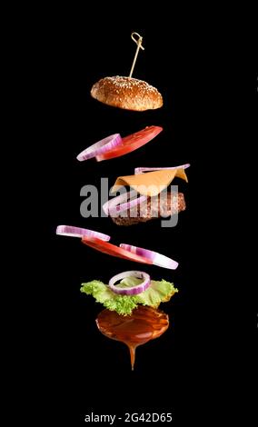 Fliegende Zutaten eines klassischen Cheeseburger Sesambrötchen, Zwiebelringe, Tomatenscheiben und einem saftigen Grillschnitzel Stockfoto