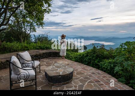 Von der Terrasse der Virunga Lodge in der Nähe von Kinyababa, Norden, blickt eine junge Frau in Sommerkleid und Sonnenhut auf den Ruhondo-See und die Berge Stockfoto