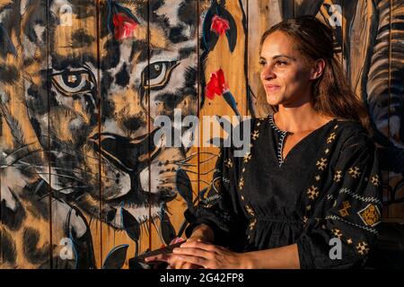 Junge Frau, die vor einem Wandbild mit Safari-Tiermotiven in einem Café, Kayonza, Ostprovinz, Ruanda, Afrika, sitzt Stockfoto