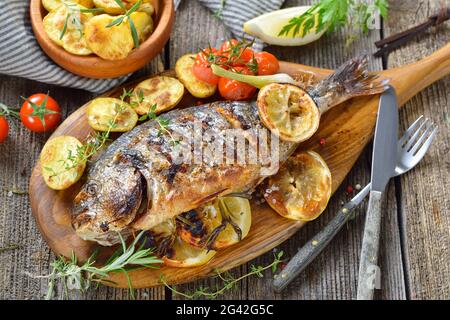 Gegrillte Goldbrasse mit Kräutern, Zitronenscheiben und Rosmarinkartoffeln auf einem ovalen Holzbrett Stockfoto