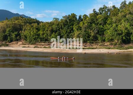 Einheimische in einem Longtail-Boot auf dem Mekong-Fluss mit Küstenlinie und üppiger Vegetation dahinter, in der Nähe von Luang Prabang, Provinz Luang Prabang, Laos, Asien Stockfoto