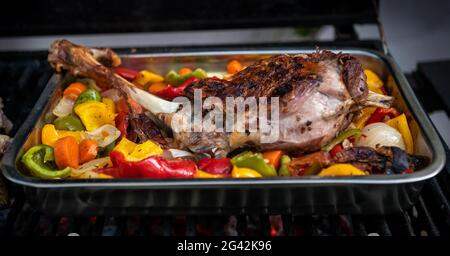 Koch kocht ein köstliches Lammkeule auf einem Grill mit farbigen Paprika und Kartoffeln in einem Metalltablett. Nahaufnahme von leckerem Ziegenfleisch auf dem Grill des Gartenhauses Stockfoto
