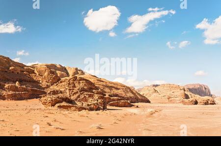 Felsige Massive auf roter Sandwüste, Fahrzeugspuren Boden, heller wolkiger Himmel im Hintergrund, typische Landschaft im Wadi Rum, Jordanien Stockfoto