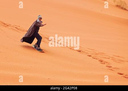 Junger Mann, der Sanddüne surft, trägt bisht - einen traditionellen Beduinenmantel. Sandsurfing ist eine der Attraktionen in der Wadi Rum Wüste Stockfoto