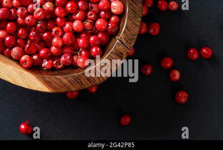 Ganze rosa/rote Pfefferkörner in kleiner Holzschale, einige verstreut auf schwarz/grauem Papier, Nahaufnahme von oben Stockfoto