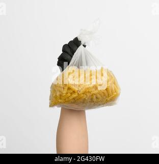 Die weibliche Hand in einem schwarzen Handschuh hält einen transparenten Beutel mit roher Fusilli-Pasta Stockfoto