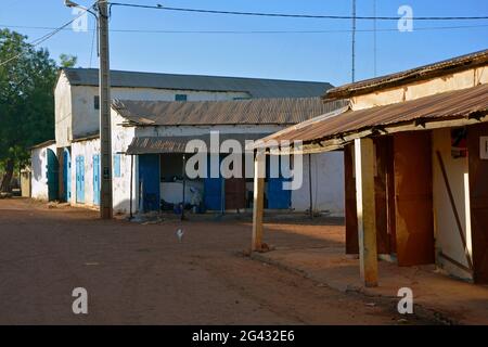 Gambia; Central River Region; Kuntaur; Hauptstraße mit Geschäften; ein Mann sitzt in seinem Laden im hinteren Gebäude; Huhn läuft auf der Straße Stockfoto