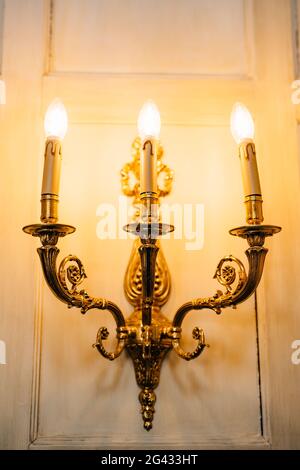 Goldene Stehleuchten mit drei Lampen, verziert mit Blumenmustern an der Wand. Inneneinrichtung einer alten Villa in Italien. Stockfoto