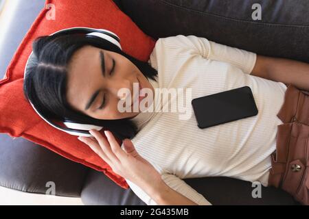 Lächelnder Mann mit gemischter Rasse und flüssigem Geschlecht, der auf der Couch liegt und trägt Kabellose Kopfhörer über das Smartphone Stockfoto