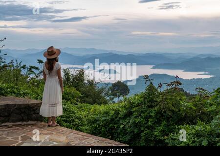 Von der Terrasse der Virunga Lodge in der Nähe von Kinyababa, Norden, blickt eine junge Frau in Sommerkleid und Sonnenhut auf den Ruhondo-See und die Berge Stockfoto