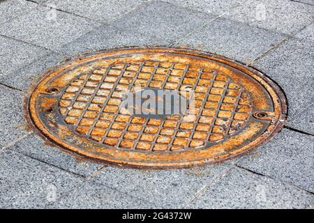 Runde eiserne rostige Luke auf einem Fußgängerweg aus grauen Steingranitfliesen in der Nähe des Kanalisationssystems der Stadt, niemand. Stockfoto