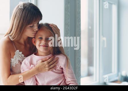 Die schöne junge Mutter und ihre charmante kleine Tochter umarmen und lächeln am Tagesfenster. Eine glückliche, liebevolle Familie, die hier übernachtet