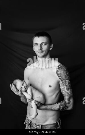 Vater mit Tätowierung am Arm hält sein neugeborenes Baby auf einem schwarz-weißen Hintergrund Stockfoto