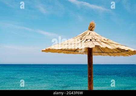 Ein Strand mit Strohschirm gegen den blauen Himmel und azurblauem Wasser an einem Sandstrand in Kroatien, in der Stadt Primosten. Stockfoto