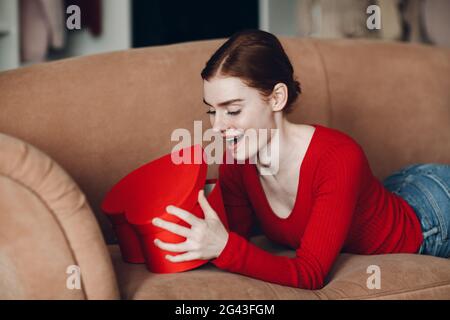 Schöne junge Frau mit roten Haaren liegt in ihrem Sofa im Wohnzimmer und hält und öffnen Sie die Geschenkbox in Form von Herz und Stockfoto