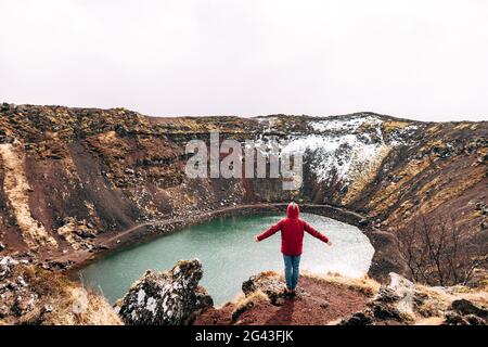 Ein Typ in einer roten Jacke steht auf einem Felsen über dem Lake Kerid - einem Krater vulkanischen See in Island. Roter vulkanischer Boden, ähnlich wie Marti Stockfoto