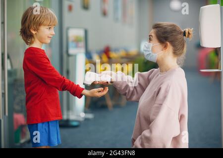 Temperaturmessung von Kindern mit einem Laserthermometer in der Grundschule. Frau in Gesichtsmaske Stockfoto