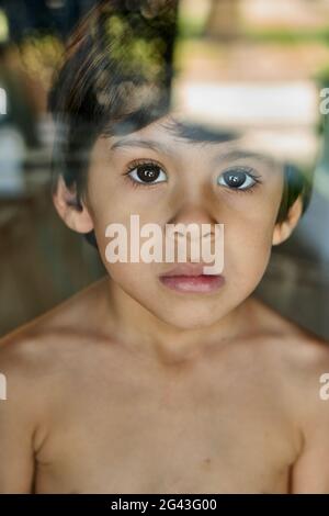 Kleiner Junge, der hinter dem Glasfenster gerade geradeaus schaut, traurig Stockfoto