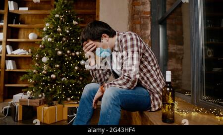 Einsamer Mann mit Covid 19 Gesichtsmaske, sehr verärgert und betrunken, sitzt am Fenster und am Weihnachtsbaum und trinkt Alkohol aus der Flasche Stockfoto