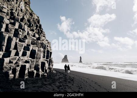 Reiseziel Island Hochzeit. Ein Hochzeitspaar spaziert entlang des schwarzen Sandstrandes von Vik, in der Nähe des Basaltfelsen, in Form von Pil Stockfoto